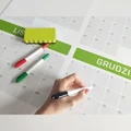 Suchościeralna tablica z nadrukiem kalendarza z zielonymi nagłówkami, akcesoriami i dłonią piszącą markerem