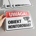 Naklejka BHP Uwaga obiekt monitorowany z czerwoną belką i kamerą, pokazana jako elastyczny arkusz w dłoni