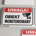 Naklejki BHP Uwaga obiekt monitorowany z czerwoną belką i czarną kamerą, ułożone w stos