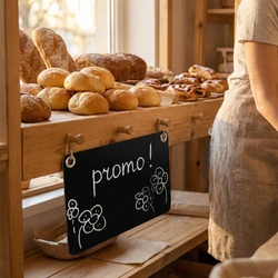 Tabliczka kredowa A4 z przykładowym napisem promo i otworami montażowymi, zawieszona na drewnianym regale w piekarni